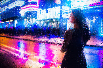 iew through glass window with rain drops on blurred reflection silhouette of a girl on a city street after rain and colorful neon bokeh city lights, night street scene. Focus on raindrops on glass	