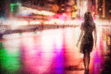 View through glass window with rain drops on blurred reflection silhouette of a girl on a city street after rain and colorful neon bokeh city lights, night street scene. Focus on raindrops on glass