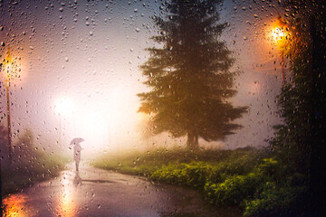 View through a glass window with raindrops on the silhouette of a man walking under an umbrella down the street in the morning mist in the rain. lights of street lamps. Focus on the raindro