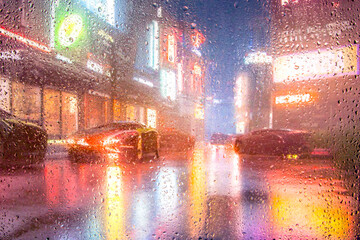 View through glass window with rain drops on blurred  city street after rain and colorful neon bokeh city lights, night street scene. Focus on raindrops on glass
