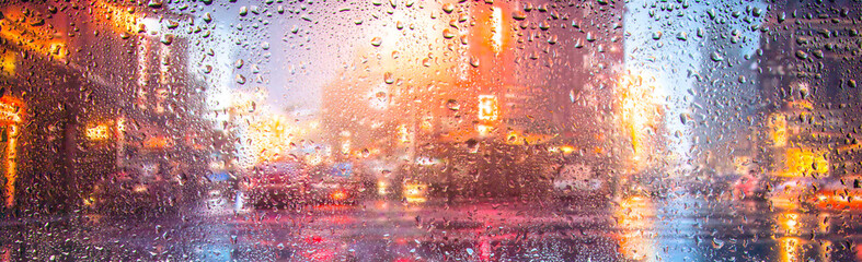 View through a glass window with raindrops on city streets with cars in the rain, bokeh of colorful city lights, night street scene. Focus on raindrops on glass