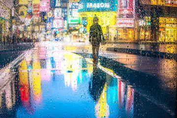 View through a glass window with raindrops on a blurred silhouette of a girl on a city street after rain against the bokeh of colorful city lights, night street scene. Focus on raindrops on glass