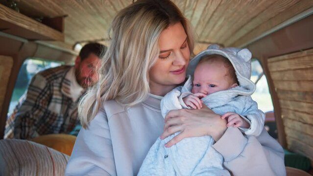 Portrait of young caucasian mother holding newborn child, kissing head, smiling. Close-up, camera-looking, camper van interior. Happy family at countryside picnic, heartwarming memories