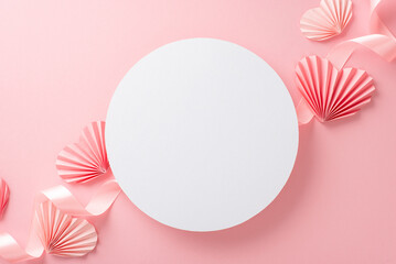 Top view photo of empty white circle with a pastel pink ribbon and paper origami hearts on an isolated light pink background with copy-space