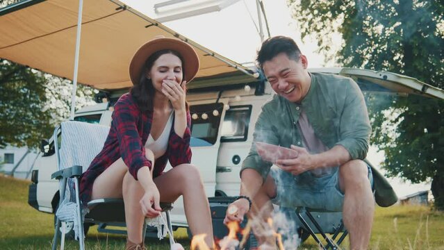 Caucasian young hipster girl and asian man sitting by campfire, roasting marshmallows, smiling, laughing. Joyful conversation at countryside picnic, pair in love having fun