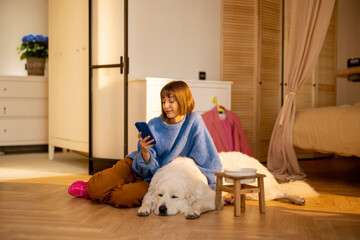 Young woman relaxes, sitting with her dog and using phone at modern cozy living room at home. Domestic lifestyle and pets concept