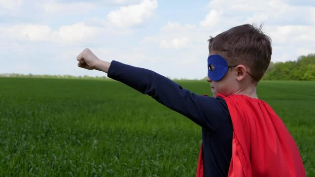 A kid in a superhero mask and cape raises his hand up, demonstrating strength and readiness to act against the background of a green meadow. Character. kids play in garden