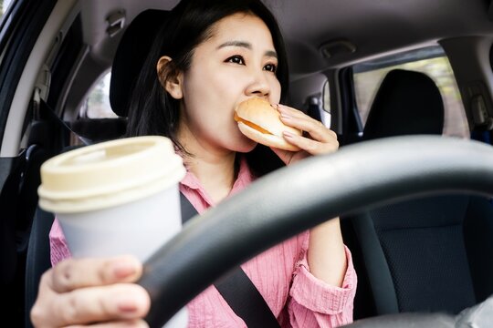 risky behavior driving concept with  Asian woman eating burger and drinking coffee while driving a vehicle, poses accident risk and traffic crashes
