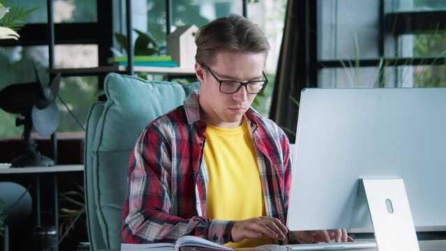 Young caucasian male working at the desktop computer, looking at the book on a table. Typing on keyboard, writing code. Finding a solution for a tricky task