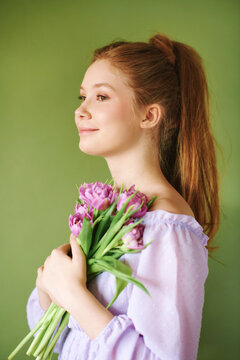 Beauty portrait of pretty young 15 - 16 year old redhaired teeenage girl wearing purple dress posing on green background, holding tulip flowers
