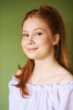 Beauty portrait of pretty young 15 - 16 year old red-haired teeenage girl wearing purple dress posing on green background
