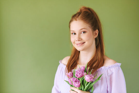 Beauty portrait of pretty young 15 - 16 year old redhaired teeenage girl wearing purple dress posing on green background, holding tulip flowers