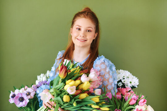 Portrait of pretty young 15 - 16 year old red-haired girl surrounded by many spring flowers posing on green background, beauty and fashion concept