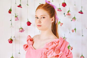 Studio portrait of pretty young teenage 15 - 16 year old red-haired girl wearing pink coral dress, posing on white background with hanging flowers, beauty and fashion concept