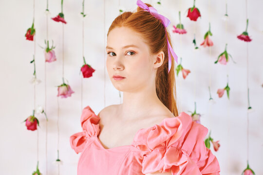 Studio portrait of pretty young teenage 15 - 16 year old red-haired girl wearing pink coral dress, posing on white background with hanging flowers, beauty and fashion concept