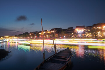 Fototapeta premium Light trails of boats with traditional lanterns on the motion. Night scene of water canal and waterfront in ancient city Hoi An in Vietnam..