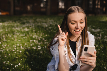 Photo portrait of blonde woman having eureka moment clarity holding phone in one hand pointing finger up sitting in park in city street. Woman wear white shirt have a good idea.