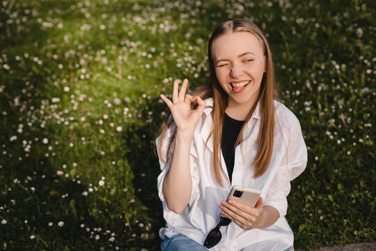 Smiling young woman hold mobile phone and show ok sign, making all right hand gesture sitting on the green fields with flowers in park. Agreement, approval, confirm. Blonde woman wink eye, show tongue