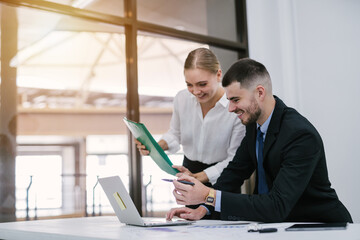 Attractive handsome white businessman sitting at desk with co-worker female holding file document consultant working on laptop computer in office.