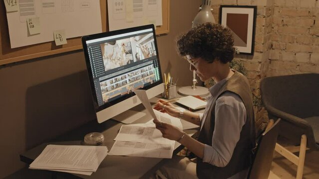 Medium shot of female Caucasian editor sitting at desk in office, studying project documents and writing notes on them
