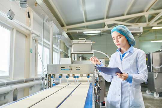 Concentrated beautiful young female food manufacturing engineer in sterile clothes analyzing data while examining industrial machine at factory