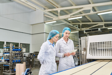 Pensive concentrated food quality engineers in sterile clothes looking at moving conveyor belt with...