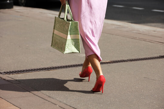 Legs of woman wearing long pink dress and red shoes on high heels walking with handbag by summer street. City female fashion in hot weather
