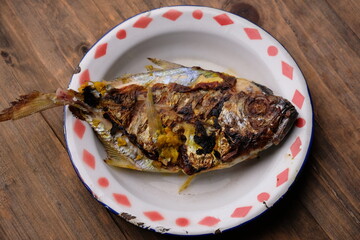 grilled pomfret on an old tin plate, on a brown wooden table. Ikan bawal bakar. 