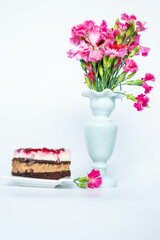 Chocolate and vanilla cake with strawberry jam near a white vase with pink carnations on a white background
