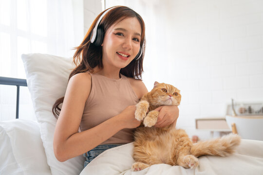 Lovely Asian girl with headphone hold and cudding orange cat and sit on bed also look at camera with smiling and day light.