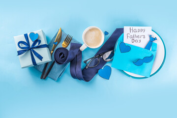 Accessory and decor arranged Father's Day greeting card, Dad`s holiday flat lay, table setting with envelope, postcard, mustaches, napkin, tie, coffee and gift box on blue background