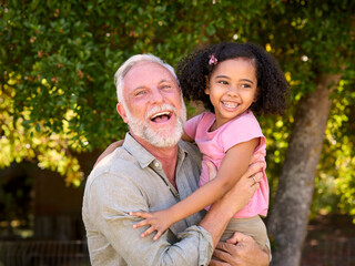 Portrait Of Multi-Generation Family With Grandfather Carrying Smiling Granddaughter In Garden