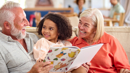 Granddaughter Reading With Grandparents On Sofa At Home With Parents In Background