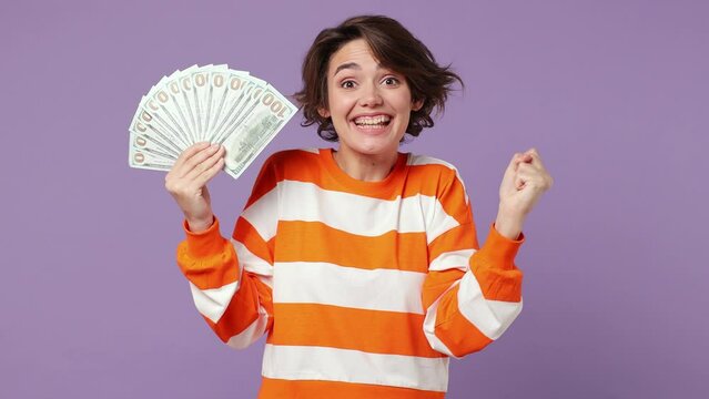 Happy fun young woman wear casual clothes hold in hand fan of cash money in dollar banknotes doing winner gesture celebrate isolated on plain pastel light purple background. People lifestyle concept