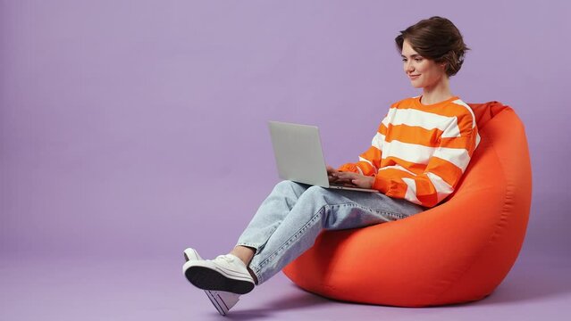 Full body caucasian young student fun smart IT woman wear casual clothes sit in bag chair hold use work on laptop pc computer isolated on plain pastel light purple background. People lifestyle concept