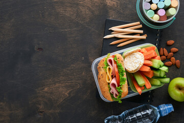 Healthy food for school lunch concept. Healthy bread, meat sandwich with cheese, apple, fresh cucumber, carrots, nuts in container, water and yogurt on dark background. Back to school concept.