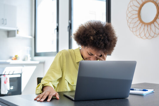 Stressed tired young Black freelance woman suffering from migraine, headache at workplace. African employee, home worker, businesswoman sitting at laptop, touching temples, holding head..