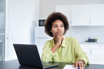 Serious young African business woman, sitting at office workplace with laptop, looking away with...