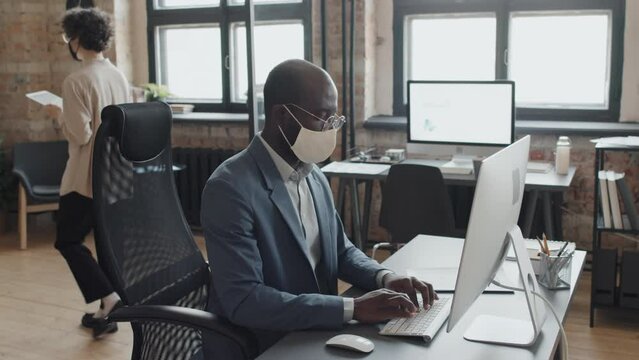 Medium tilt shot of male African American manager wearing mask and working in computer while young Caucasian woman in mask going along office with tablet in hands