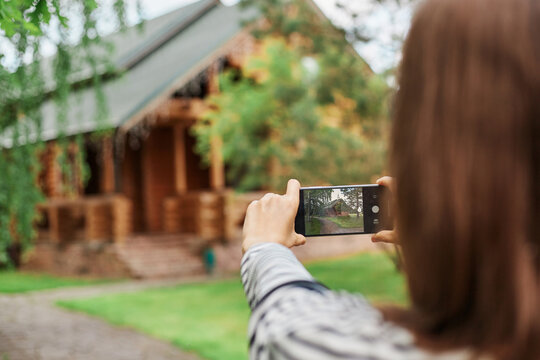 Back view of brown haired woman photographing wooden house in part vising new places sightseeing traveling having vacation shooting video for her travel blog.