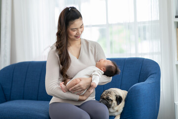 Beautiful New Asian mom holding newborn baby in her arms sit on couch with cute dog pug breed at cozy home.Happy infant baby sleep in mother arms safety and comfortable.Mom and Baby Concept