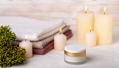 Face cream and burning candles, stack of towels and juniper branch on light background. Concept of calmness, comfort, spa treatments. Selective focus