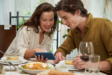 Obraz na płótnie Canvas Cheerful young woman showing photo on smartphone to boyfriend when they are eating dinner at home