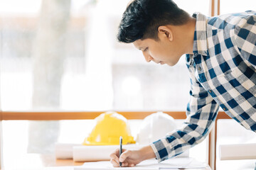 A side view of an Asian male architect looking smart, like an engineer, drawing a blueprint with a pen. Concept of industrial project design in office. Wearing suit, helmet on desk.