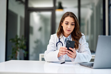 A serious female doctor using a mobile phone while sitting at the office table.