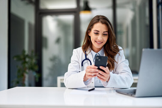 A Female Doctor Sitting At The Office Table In Front Of The Laptop And Using A Mobile Phone.