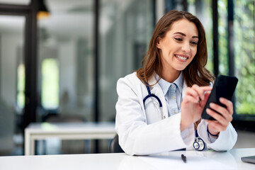 A smiling female doctor using a mobile phone while sitting at the office. - 608341462