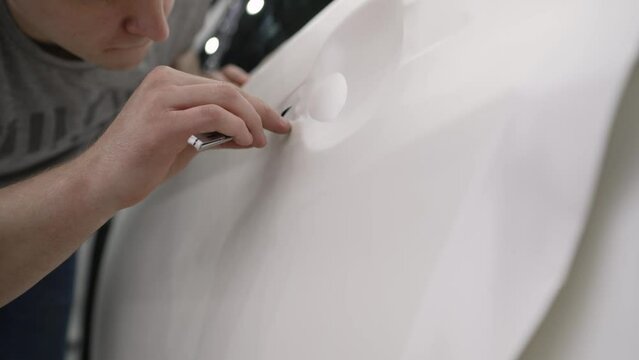 A man cuts the protective film off the door with a knife. Application of a protective film on a car door. A male specialist cuts a protective vinyl wrap on a car body for a handle in a car service