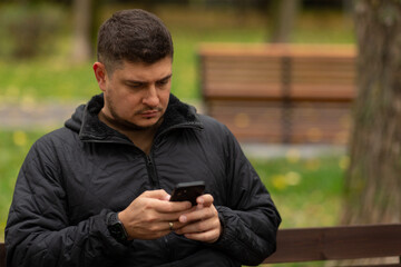 A man is sitting on a bench in the park in autumn, typing a message in his smartphone