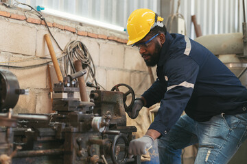 Male factory worker working at work maintenance machine in industrial factory while wearing safety uniform, glasses and hard hat. Black male technician and heavy steel lathe machine in workshop